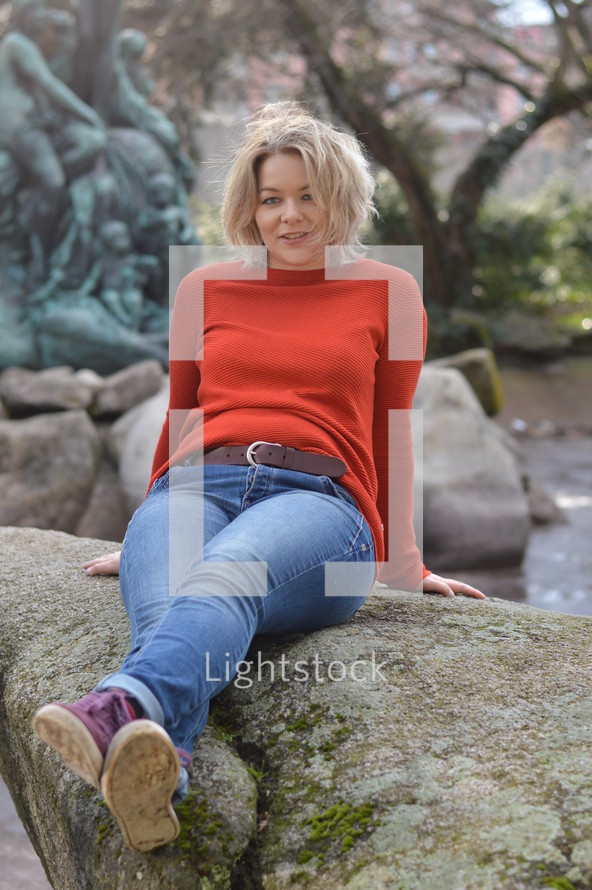 young pretty blond woman with orange pullover sitting outdoors in a park on a mossy rock smiling