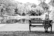 Girl sitting on a park bench by a lake surrounded by trees.