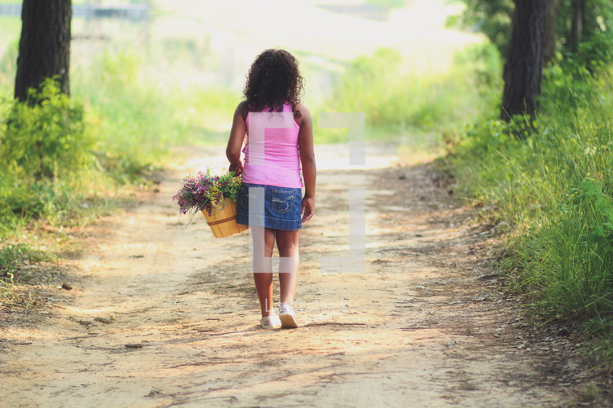 a girl carrying a basket of flowers down a dirt road 