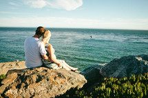 couple sitting on rocks near a beach looking out at the ocean 