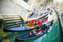Two gondolas dock by a street in a waterway between buildings
