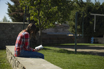 Young woman sitting outside on rock wall reading her Bible