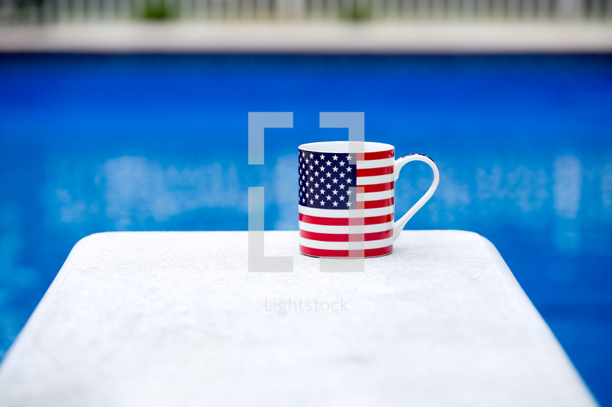 A flag coffee mug on a white table next to a blue swimming pool.