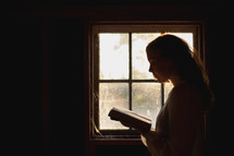 a woman reading in front of a window in a dark room 