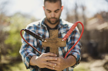 Man with eyes closed and head bowed holding cross with jumper cables attached.