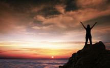 silhouette of a woman standing on a rock in front of the ocean at sunset with her hands raised in worship to God