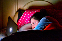 teen girl looking at a computer screen laying in bed 