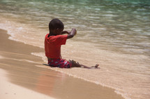 a child sitting on a beach letting the tide roll over his legs 