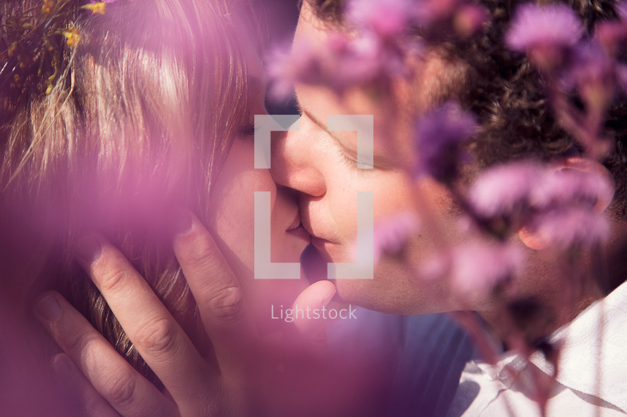 Man and woman kissing behind purple flowers.