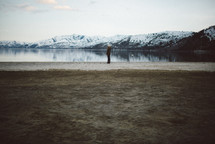woman standing by a lake surrounded by snow capped mountains 