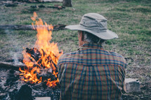 A man sit next to a camp fire.