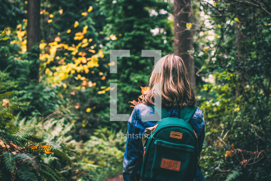 a girl with a backpack walking into the woods 