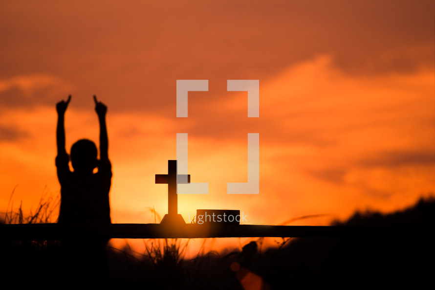 silhouette boy with hands raised and cross at sunset 