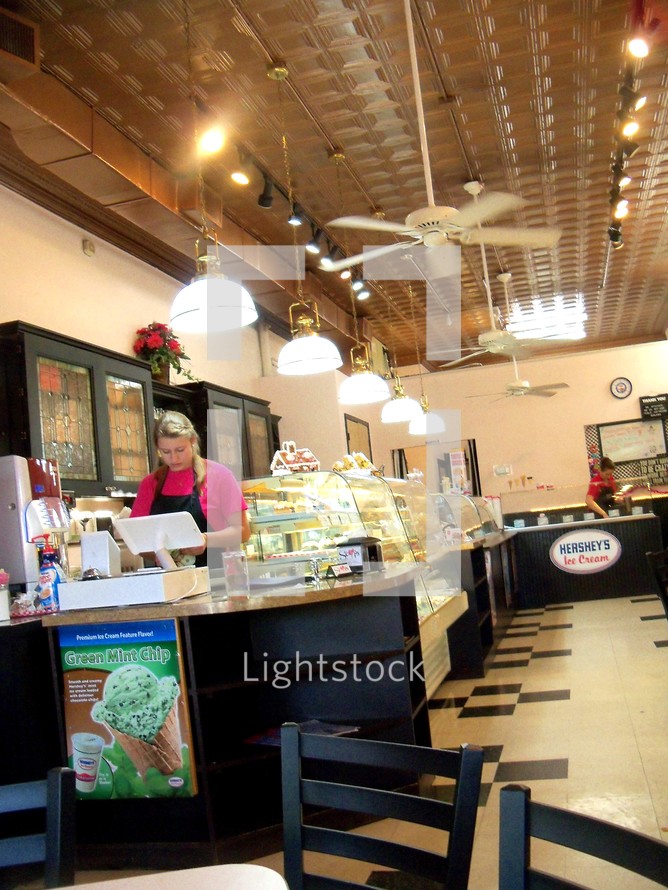 A blonde-haired female rings up a sale at a cash register while working in a small business ice cream parlour located in a small town. 