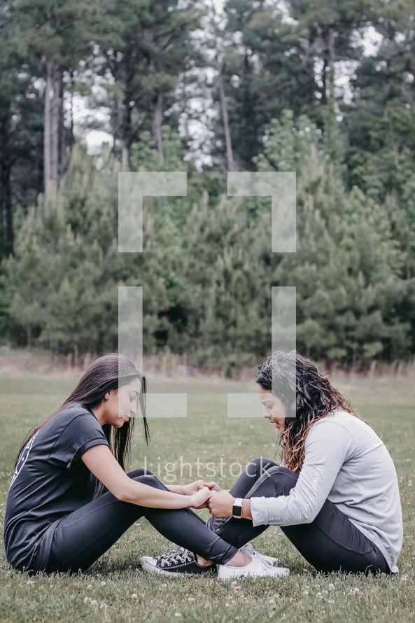 two women sitting in grass holding hands and praying 