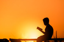 a boy sitting reading a Bible at sunset 