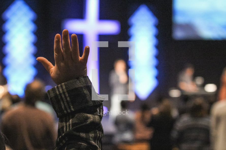 raised hand during a worship service with a glowing cross in the background 