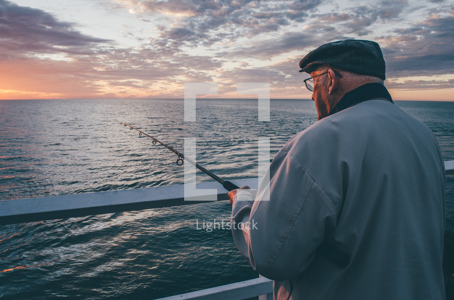 An older man fishes from a pier at sunset