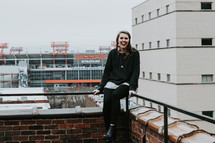 a smiling woman sitting on a railing on a roof in a city 