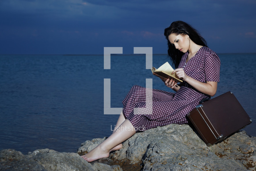 a woman reading a book on a rock by water with a suitcase 