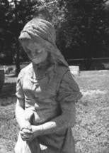 A stone statue of a weeping woman watching over a grave in a cemetery filled with tomb stones of those dearly departed.
