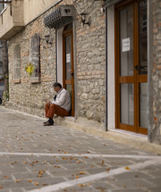 man sitting alone outside a shop along a rock wall