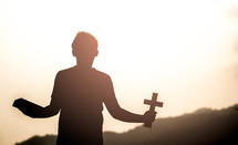 a silhouette of a boy holding a cross and Bible at sunrise 