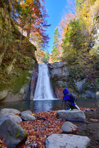 kid near photo camera on tripod with a waterfall in background in autumn