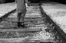 a boy in rain-boots standing on railroad tracks 