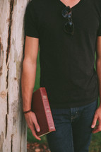 torso of a man leaning against a tree holding a Bible 