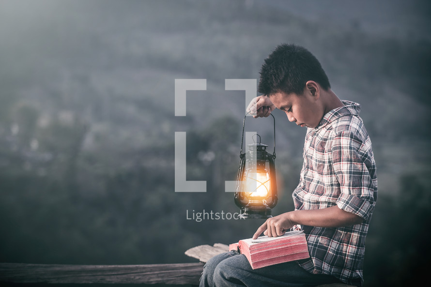 a boy reading a Bible outdoors holding a lantern 