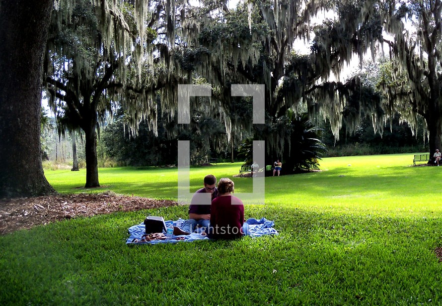 A young man and woman relax on a sunny afternoon at a picnic in a green grassy field surrounded by trees on a fall day. 