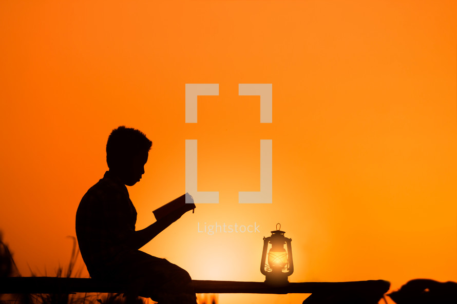 silhouette of a boy reading holy bible with Oil lamp on wood at sunset against an orange sky 