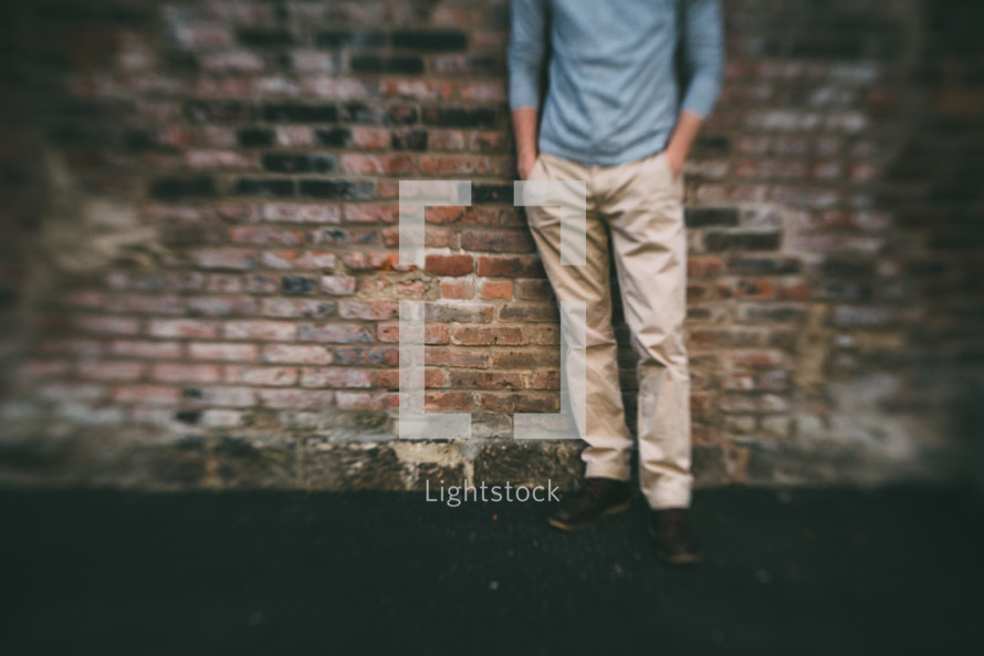 a man with his hands in his pockets leaning against a brick wall 