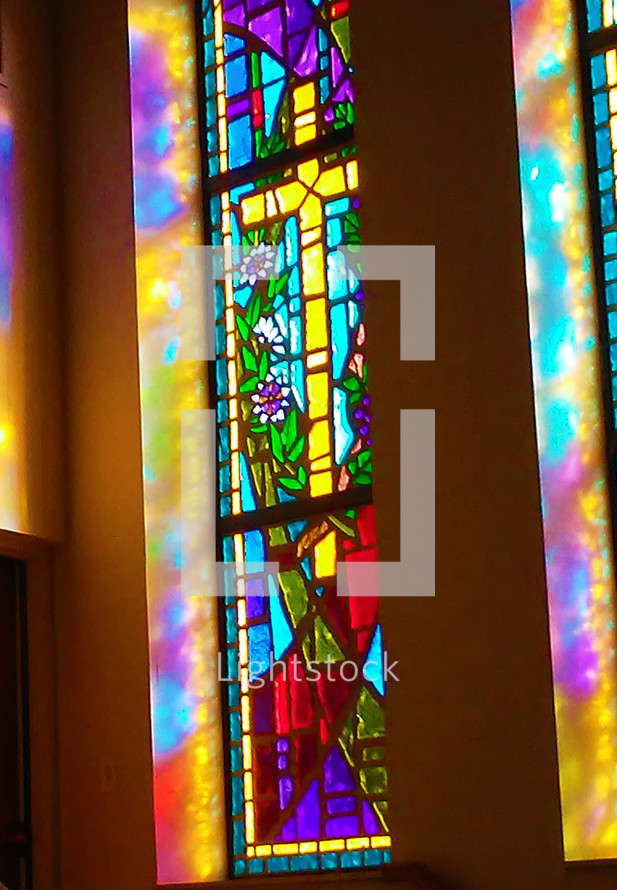 Morning sunlight and sun rays pour through a beautiful and colorful stained glass window showing a light prism of colors through a large stained glass window of the cross in the morning sunlit church service. 