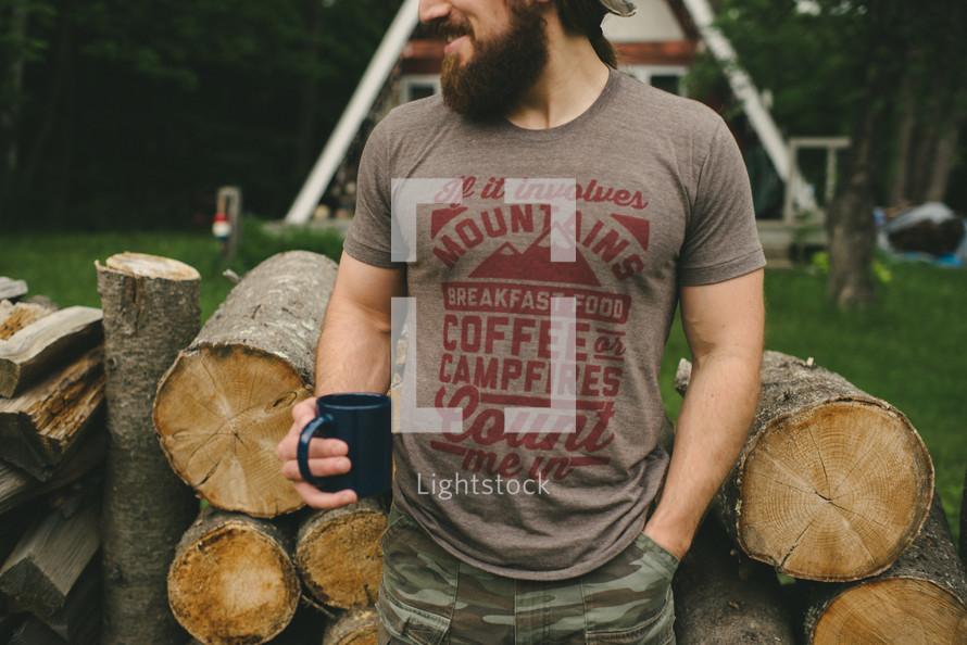 A bearded man in a camping t-shirt stands before a pile of firewood.