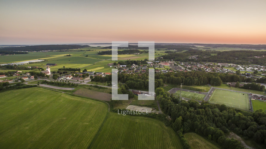 Aerial view of the small town Fjärås, Kungsbacka, in Sweden. Beautiful and lush farmlands with forest surrounding them while the sun sets and warms the atmosphere.