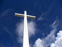A white cross towers over the earth, sky and clouds reaching out to Heaven against a blue sky on a bright sunny day giving hope to all who see it that there is a God who came down from Heaven to be with us and take our place on this very cross for our sins that we might be saved and spend eternity in Heaven with Jesus.