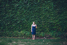 A woman standing in front of an ivy covered wall. 