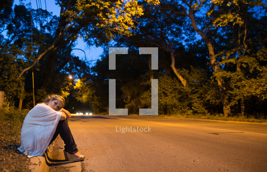 Woman sitting on the side of the road at night.