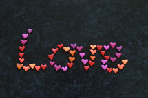 The word LOVE written with many little colorful clay hearts on black background.