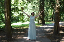 a woman in a long dress standing on a path in a forest with raised hands 