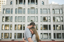 A man and woman hug in front of a large building.