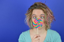 portrait of a young blond woman with colourful streaks in the hair and a big sweet multicolored lollipop in her hands squinting at it and having fun