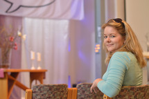 a young blond woman sitting at a chair in a modern church before the service starts turning around and looking back over her shoulder. - useful for church reopening: welcome back to church