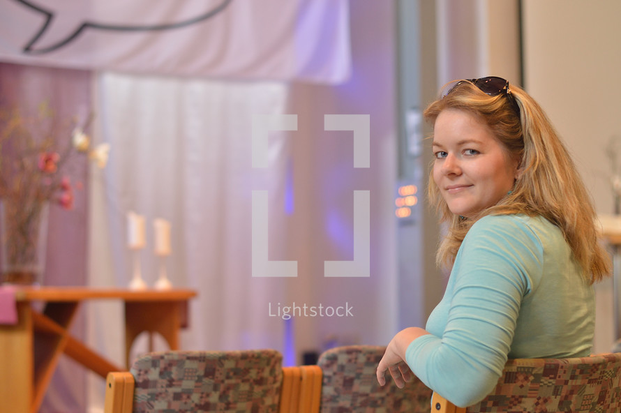 a young blond woman sitting at a chair in a modern church before the service starts turning around and looking back over her shoulder. - useful for church reopening: welcome back to church