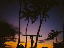 A person with a surfboard on his head is silhouetted against the sunset.