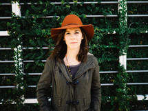 A young woman in hat and coat stands in front of an ivy covered wall.