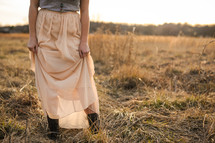 a woman in a long skirt and cowboy boots standing in dry grasses 