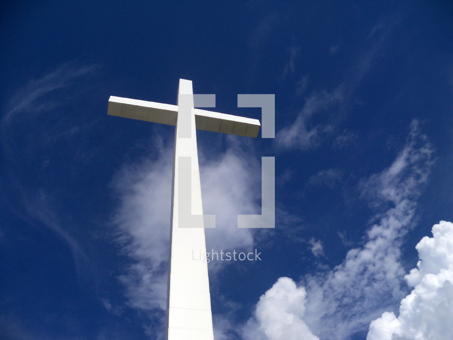 A white cross towers over the earth, sky and clouds reaching out to Heaven against a blue sky on a bright sunny day giving hope to all who see it that there is a God who came down from Heaven to be with us and take our place on this very cross for our sins that we might be saved and spend eternity in Heaven with Jesus.